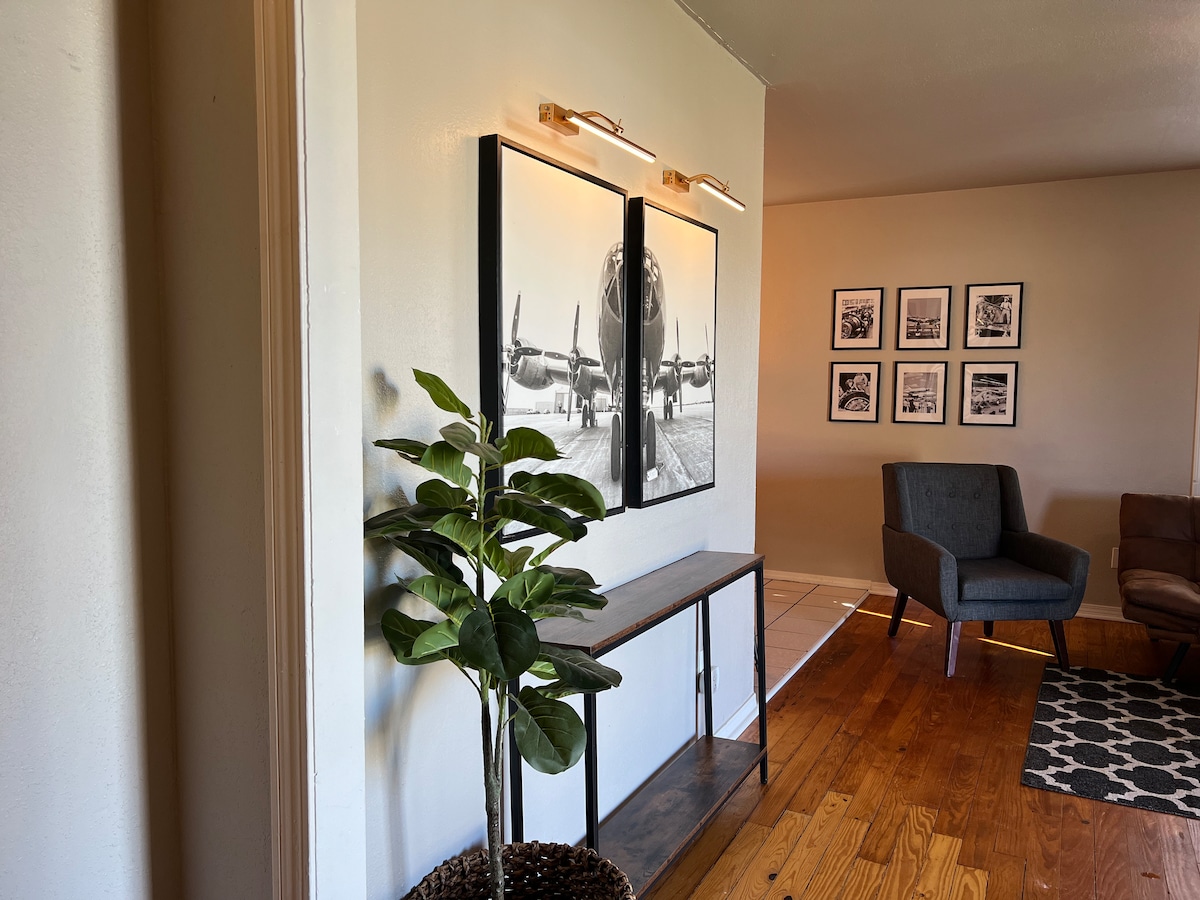 Elegante espacio de entrada con cálidos pisos de madera, una pared de arte con temática de avión con iluminación decorativa y una moderna mesa consola con una exuberante planta de interior. Conduce a una acogedora sala de estar con un sillón oscuro y una alfombra estampada.