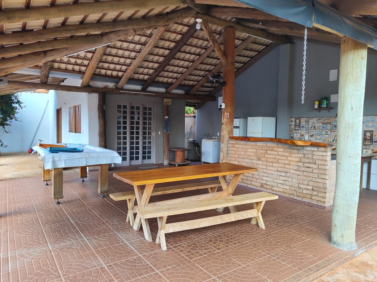 An outdoor covered space features a wooden dining table and benches, complemented by a billiard table. A rustic bar area with brick detailing is visible in the background, along with a refrigerator. The ceiling is supported by wooden beams, providing shade and comfort.