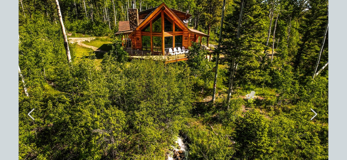 The log cabin is surrounded by lush greenery, with multiple trees framing the exterior. A deck with seating is visible, inviting relaxation amidst nature. A stream runs nearby, adding to the tranquil setting and enhancing the cabin's connection to the natural landscape.