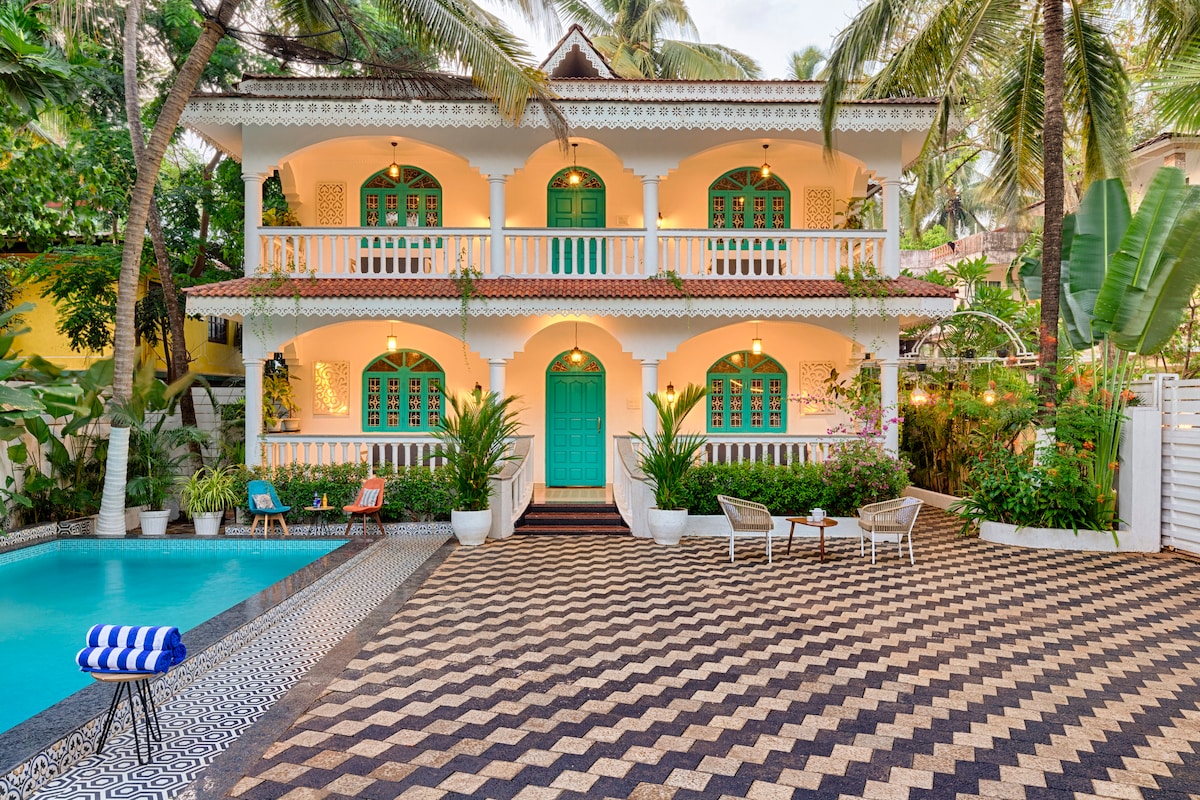 A vibrant villa is set in a lush garden, featuring an expansive pool and intricate patterned flooring. The entrance showcases striking turquoise doors framed by white arches. Comfortable seating is placed under palm trees, creating a welcoming outdoor space for relaxation.