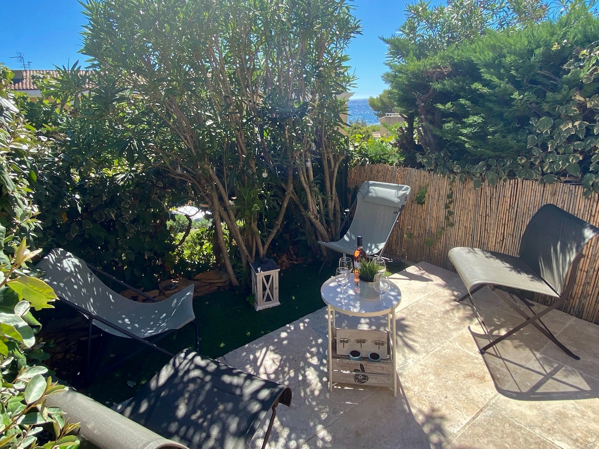 An outdoor terrace space is surrounded by lush greenery, featuring comfortable lounge chairs and a small table. The area is brightened by sunlight, creating a serene setting for relaxation. A decorative lantern adds a touch of charm to the inviting space.