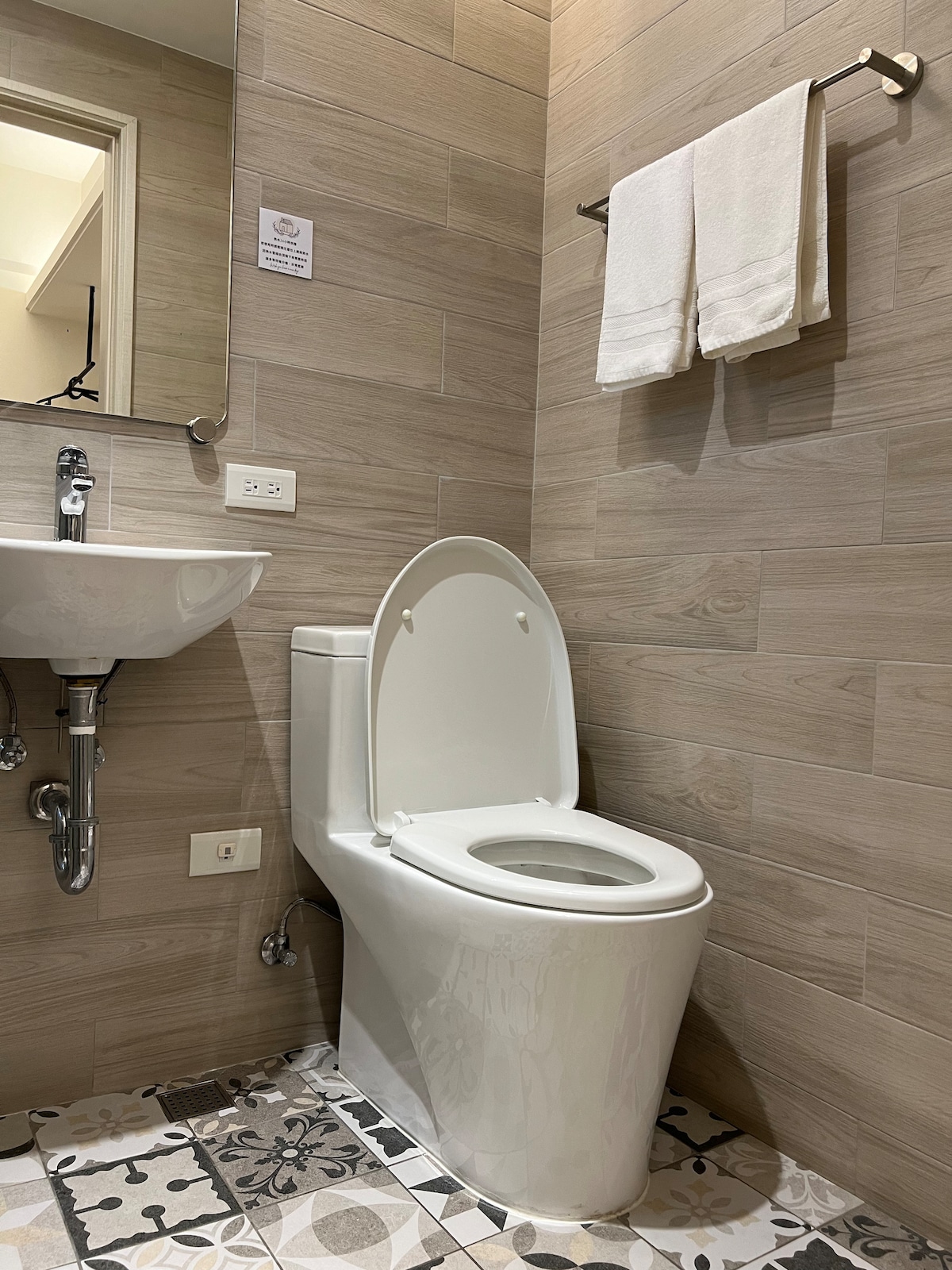 A clean and functional bathroom is visible, featuring a modern toilet with a closed lid and a wall-mounted sink. Light wood paneling enhances the space, and decorative floor tiles add a subtle pattern. Fresh towels are neatly displayed on a rack above the sink.