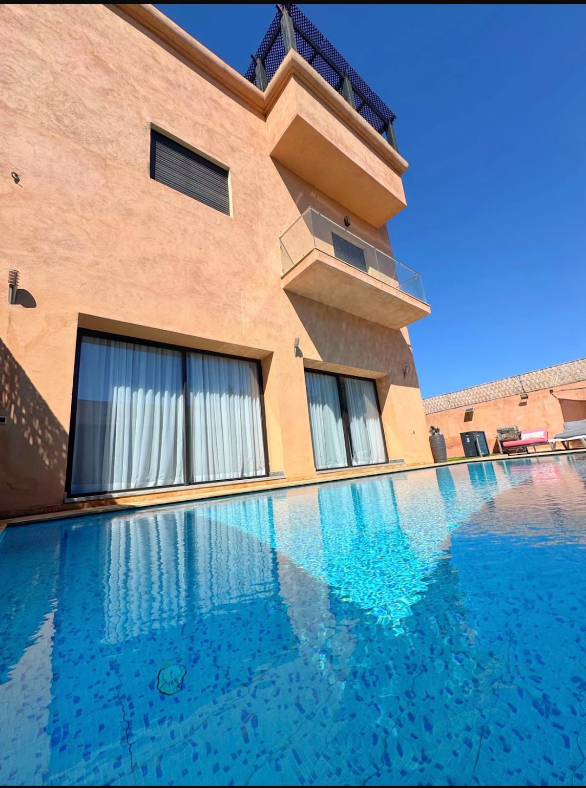 The exterior of the villa is showcased with a modern design, featuring large glass doors that open to a sparkling blue pool. The smooth surface of the water reflects the clear sky and the light-colored building structure, enhancing the serene ambiance.