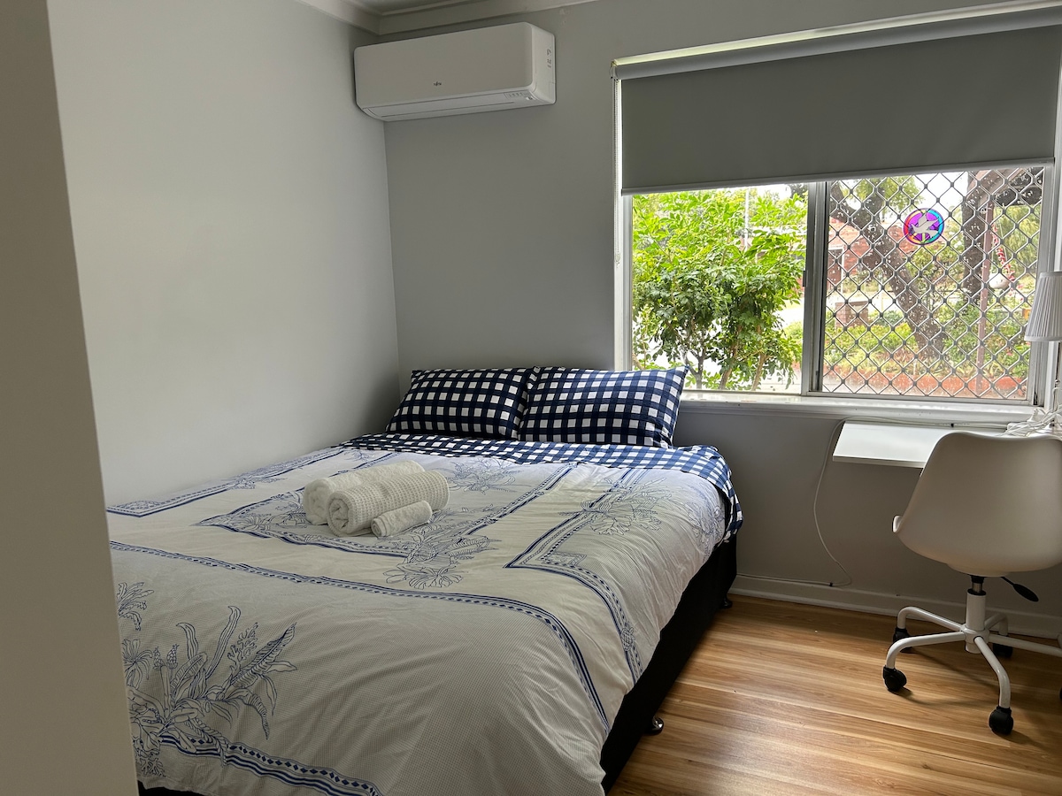 A cozy bedroom is equipped with a comfort Queen-size bed featuring a patterned duvet cover and coordinated pillows. A desk with a chair is positioned near the window, which allows natural light to illuminate the space. An air conditioning unit is mounted on the wall.