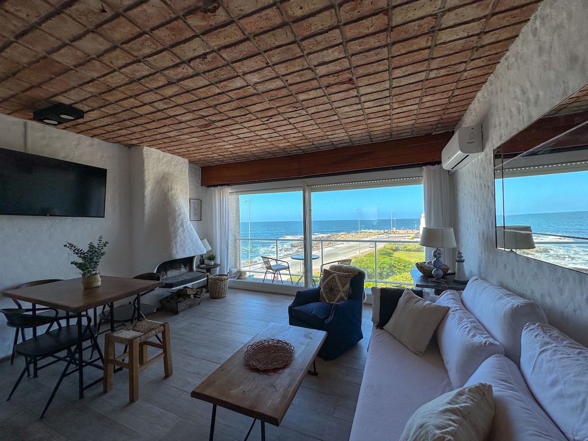 A comfortable living room features a spacious sofa and a dining table with seating. Large windows offer panoramic views of the sea and lighthouse, with natural light illuminating the area. A wall-mounted TV and air conditioning are included for convenience.