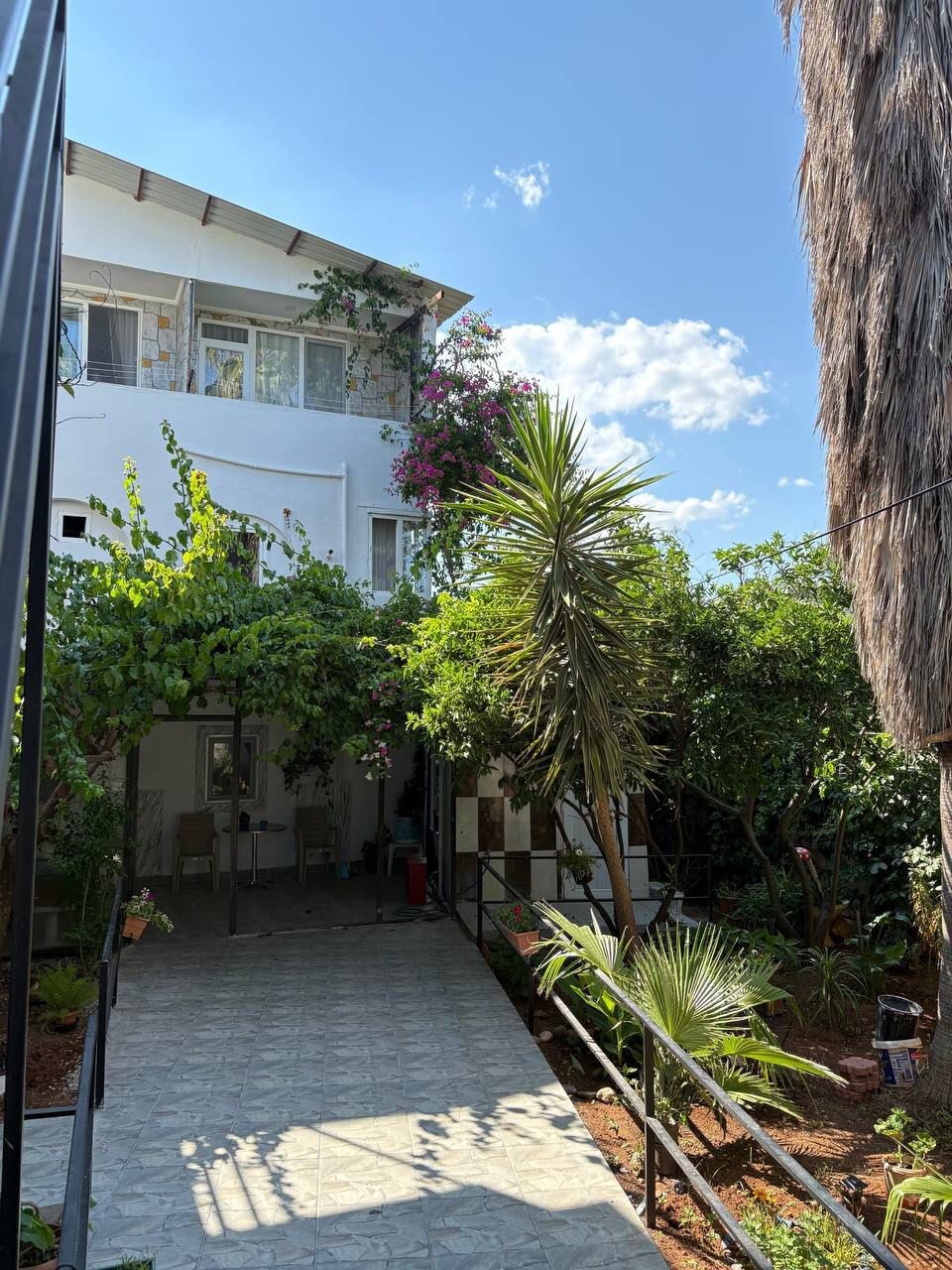 A garden path leads toward the entrance of a two-story building, surrounded by lush greenery. Tropical plants, including a tall palm, frame the walkway, which is bordered by light-colored stone tiles. Bright blue sky and scattered clouds are visible above, enhancing the natural setting.