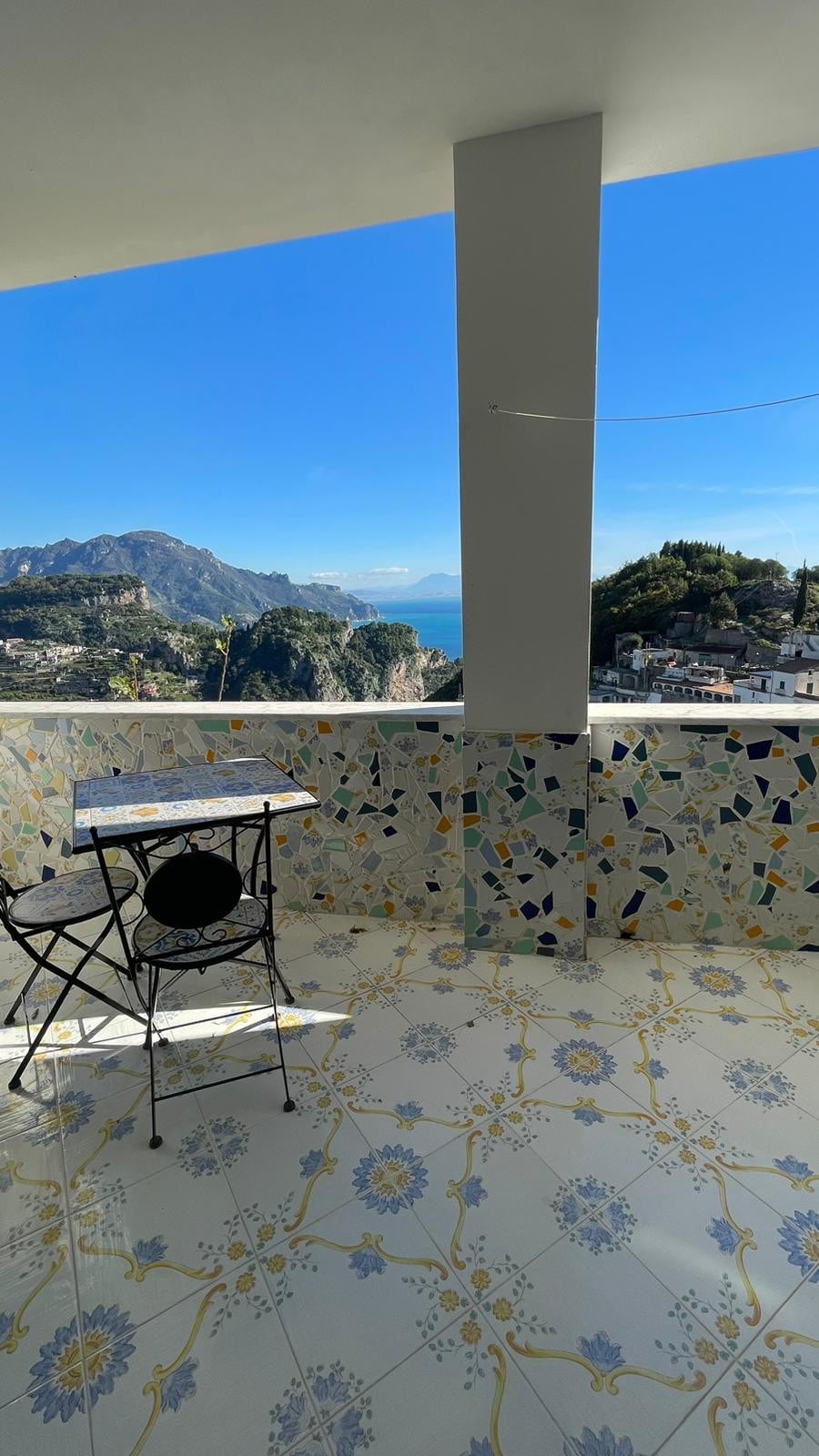 A terrace features a small table and a black chair, surrounded by colorful tiled walls. The view showcases a landscape of mountains and the sea under a clear blue sky, providing a peaceful outdoor space.