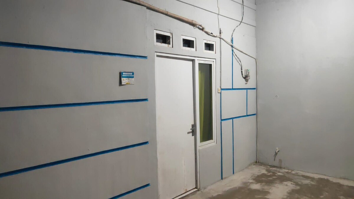 A simple interior space is depicted, featuring a solid gray wall with horizontal blue stripes. A white door is prominently visible, accompanied by three small windows arranged above it. The floor appears unfinished, enhancing the minimalist aesthetic of the area.