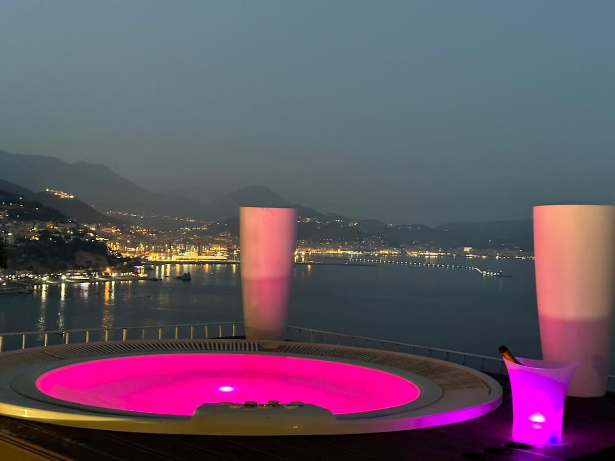 A solarium features a round hot tub illuminated in soft pink, overlooking a serene coastal view. The distant lights of a town twinkle along the shoreline, framed by mountains in the background under a twilight sky.