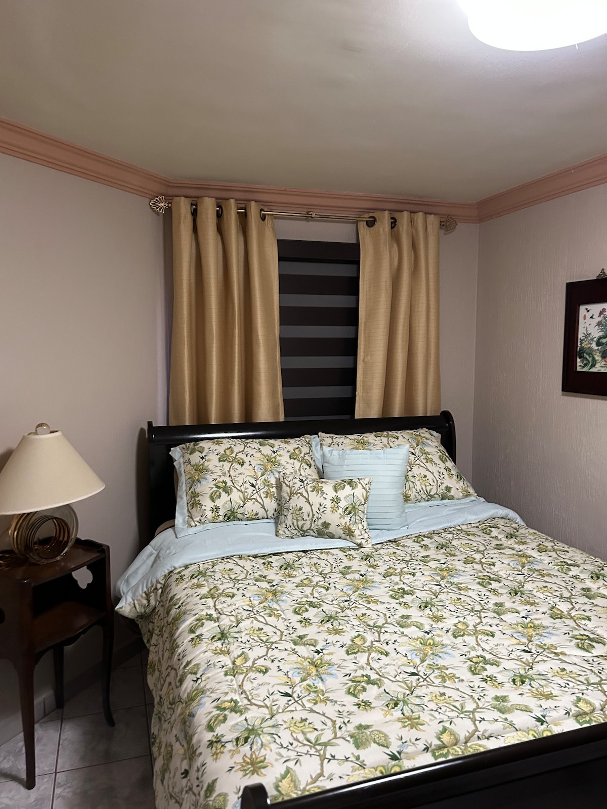 A cozy bedroom features a dark wooden bed with a floral patterned comforter and several accent pillows. Soft, light-colored curtains frame a window with horizontal blinds. A small nightstand holds a lamp, complemented by a decorative wall piece, adding to the room's welcoming atmosphere.
