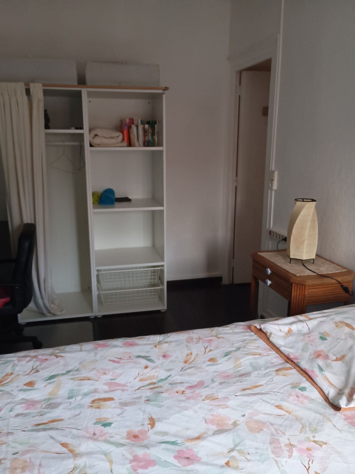 A calm bedroom space is presented, featuring a spacious bed adorned with floral bedding. A wooden nightstand with a lamp complements the room. A white shelving unit provides storage, with books and items neatly arranged. A black office chair is positioned nearby.