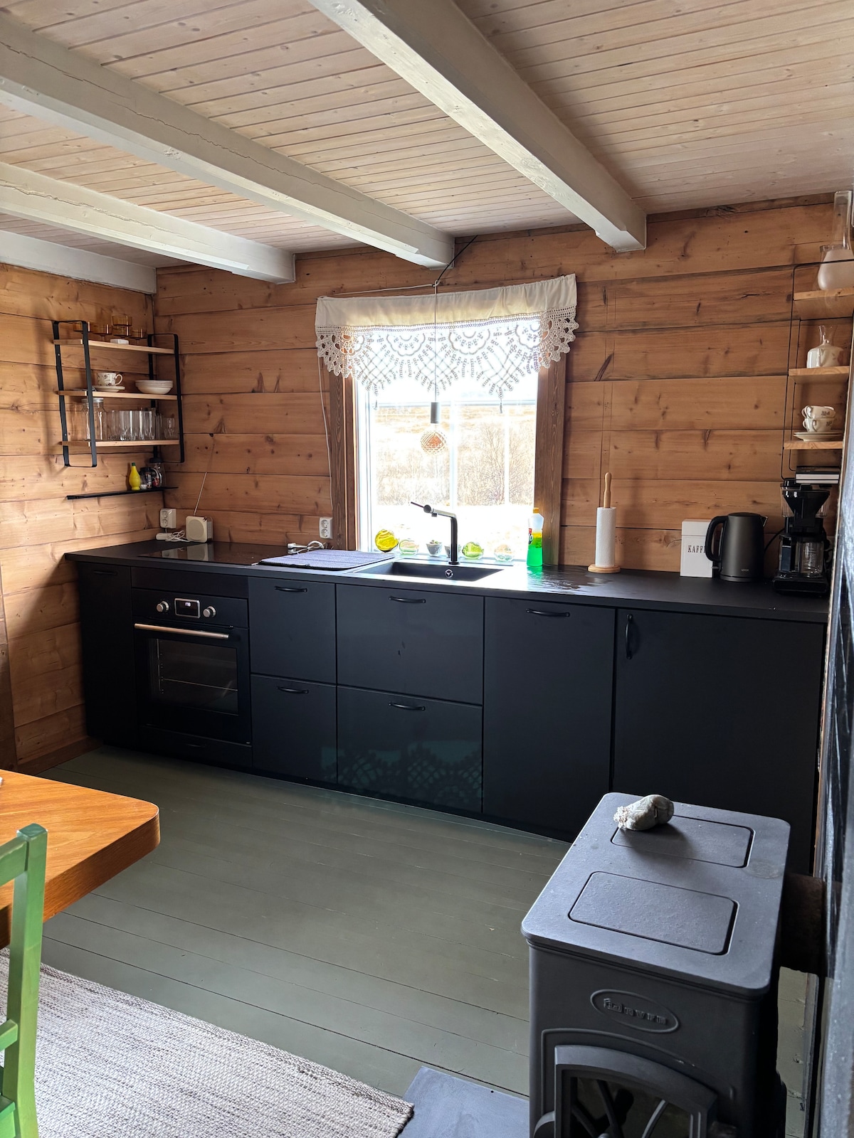 The kitchen features a blend of dark cabinetry and wooden walls, creating a warm aesthetic. A window allows natural light to illuminate the space, highlighting the compact stove and coffee maker. Shelves are arranged with dishware, while a wooden table is positioned nearby.