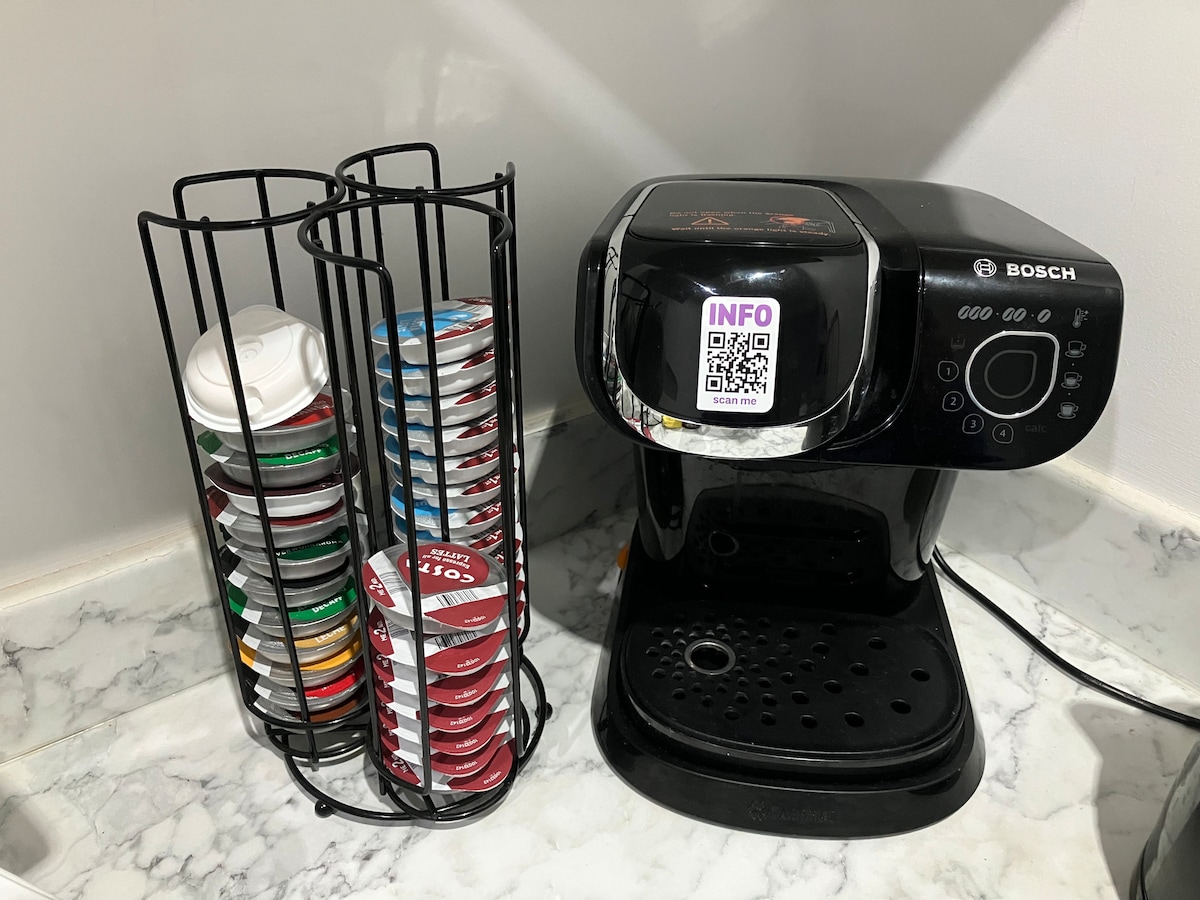 A coffee machine is positioned on a marble countertop, accompanied by a wire rack containing an assortment of coffee pods in various colors. The setup provides convenient access for preparing beverages.