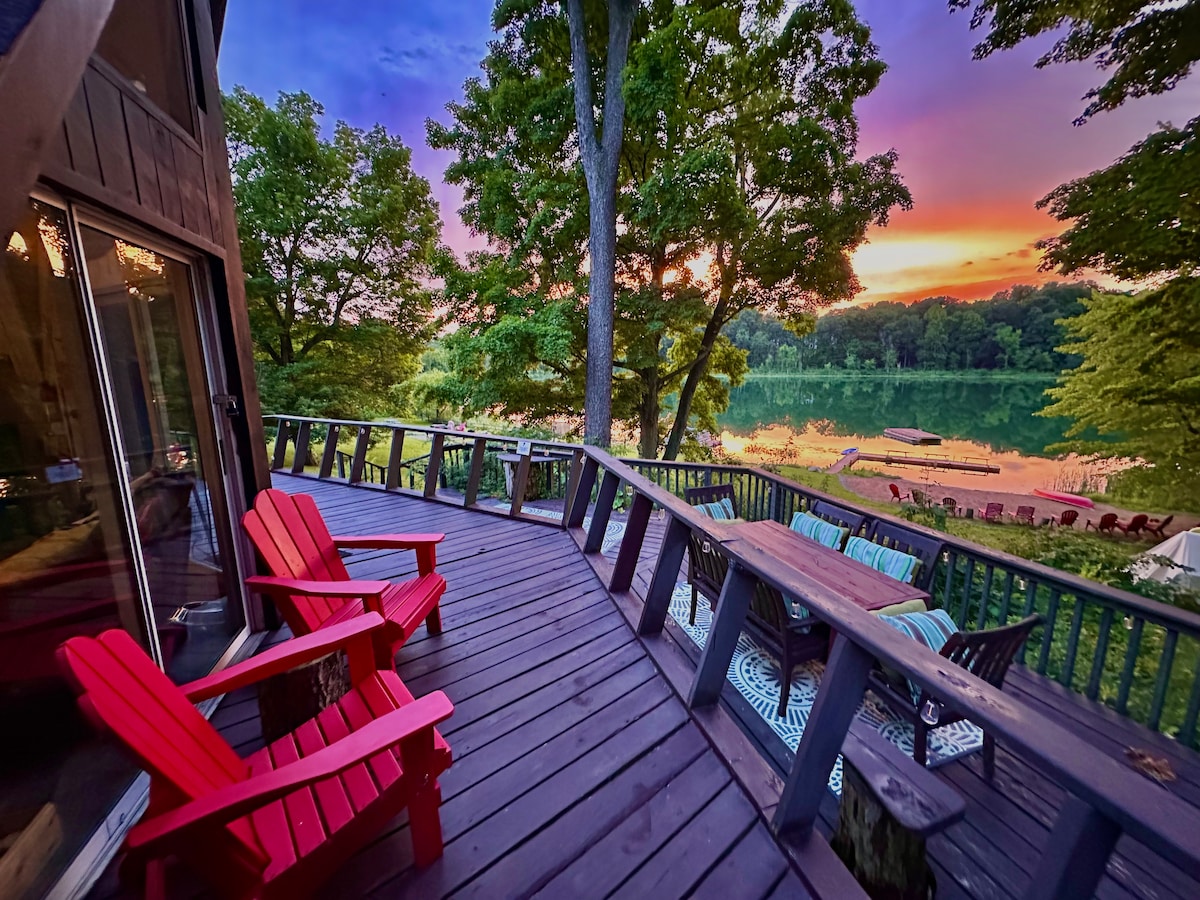 A spacious deck is viewed, featuring bright red Adirondack chairs and a wooden table surrounded by comfortable seating. The lake sparkles in the background, reflecting hues of sunset across the sky, with trees providing a lush green frame.