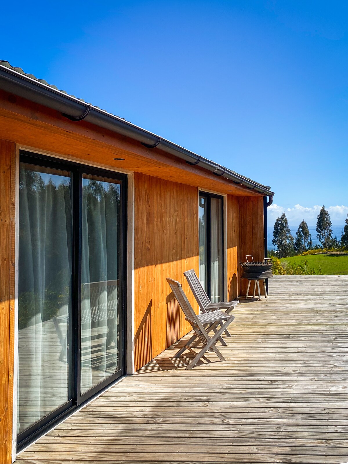 A wooden deck extends from the house, featuring two simple chairs positioned for outdoor seating. Floor-to-ceiling glass doors offer views of the surrounding landscape, while a small barbecue grill is placed nearby. The clear blue sky provides a bright backdrop.