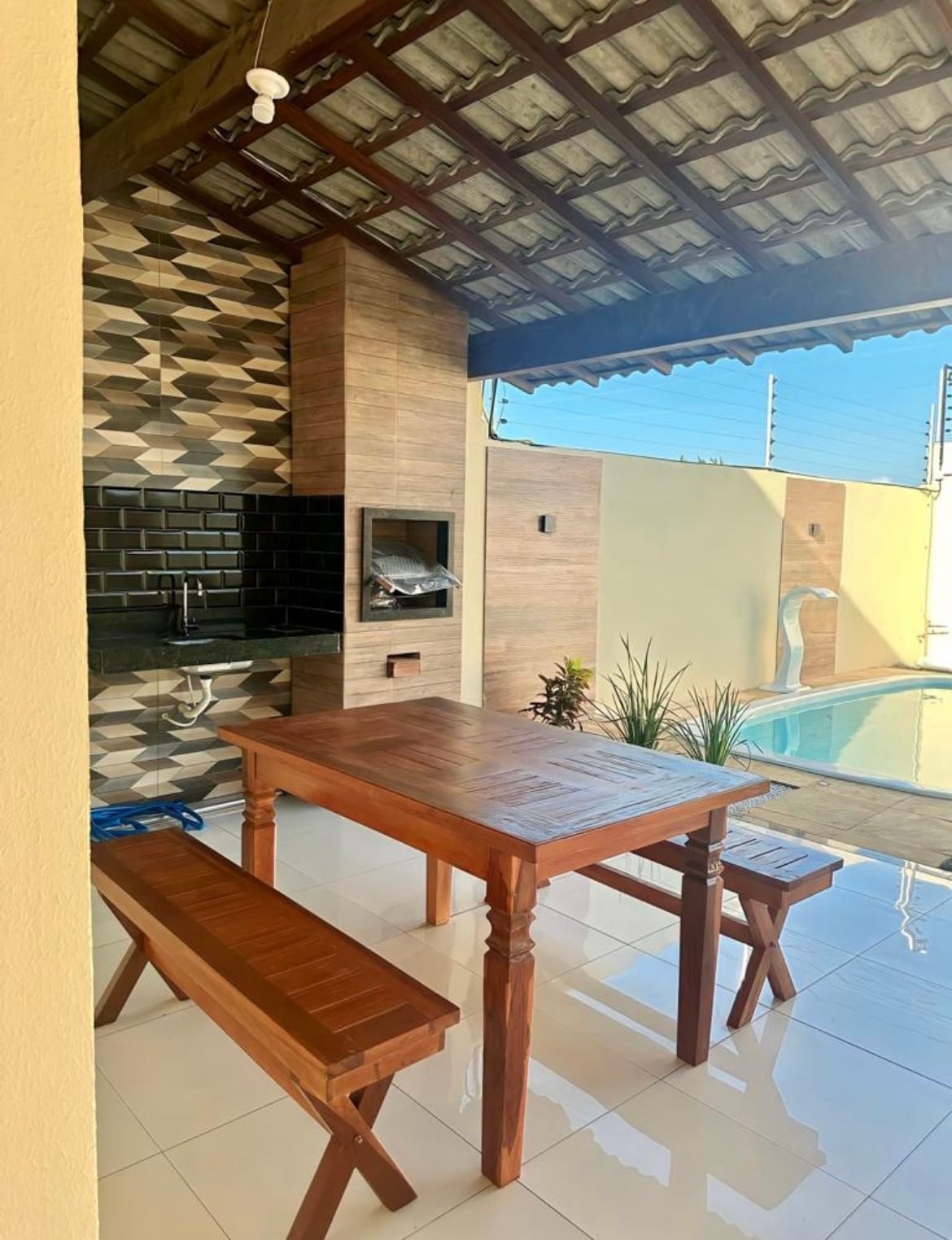 An outdoor area features a wooden dining table surrounded by two benches. A built-in barbecue grill is visible against a patterned wall. The space is complemented by a blue water pool in the background and lush greenery nearby, set under a shaded roof.