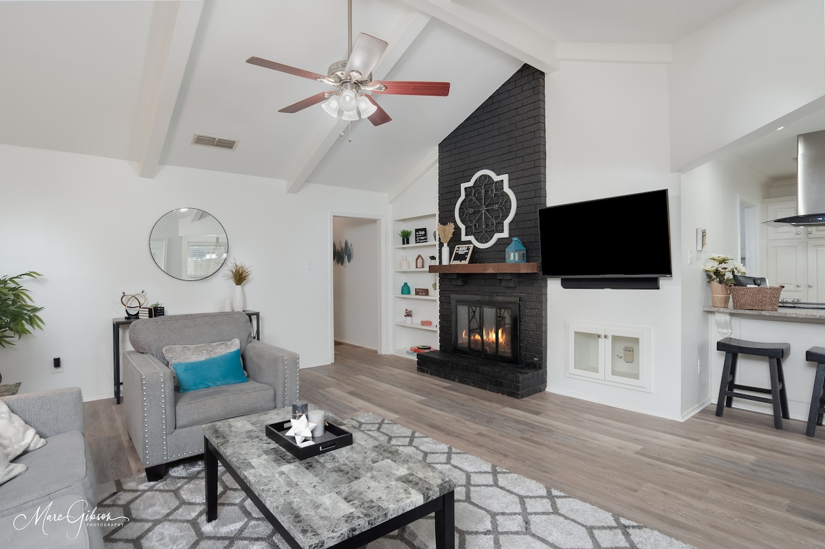 The living room features vaulted ceilings and a distinctive brick fireplace. A modern gray sofa is positioned near a coffee table, which is topped with decorative items. A flat-screen TV is mounted on the wall, complemented by natural light from nearby windows.