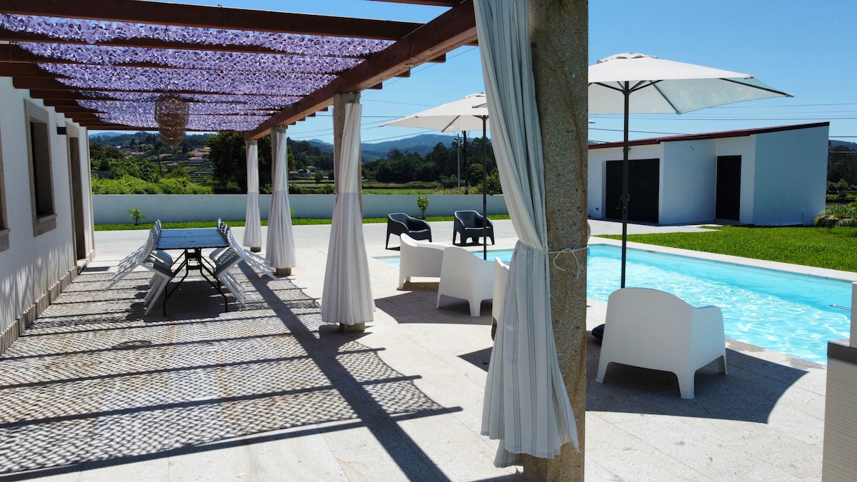 A shaded patio area features a long table with chairs, inviting relaxation beside an unobstructed view of a private pool. Umbrellas provide additional shade, while lounge chairs enhance the outdoor space. Lush greenery and a well-maintained lawn surround the area, creating a serene environment.