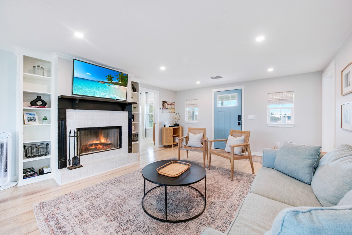 A bright living area features a modern fireplace set in a light-colored stone surround. Comfortable seating includes a sofa and two armchairs, arranged around a circular coffee table. A flat-screen television is mounted above the fireplace, complementing the welcoming atmosphere.