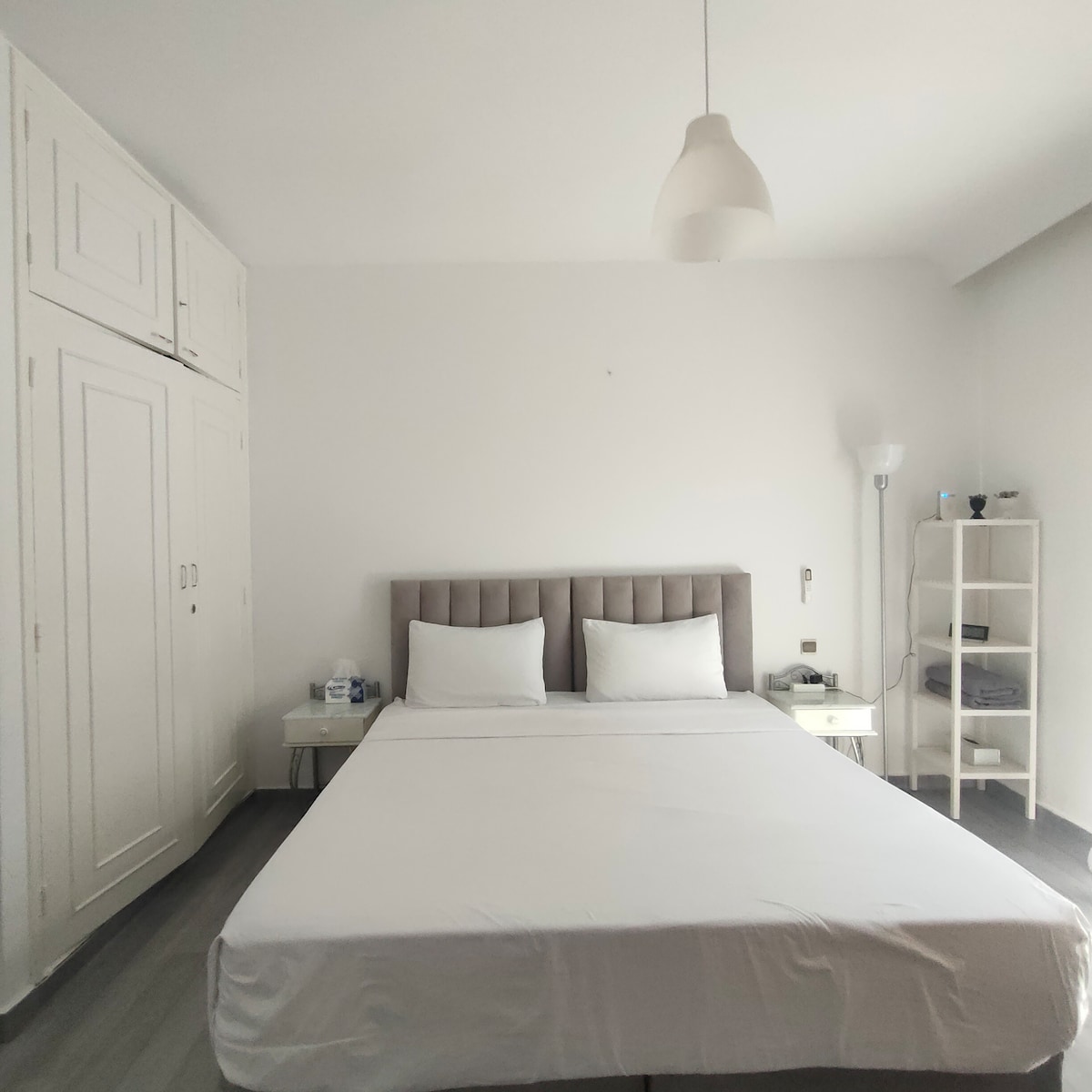 A spacious bedroom features a neatly made bed with two white pillows against a light gray headboard. Simple nightstands are positioned on either side. A tall cabinet with multiple doors provides storage, while a shelving unit holds neatly stacked towels. Soft lighting is provided by a pendant light and a floor lamp.
