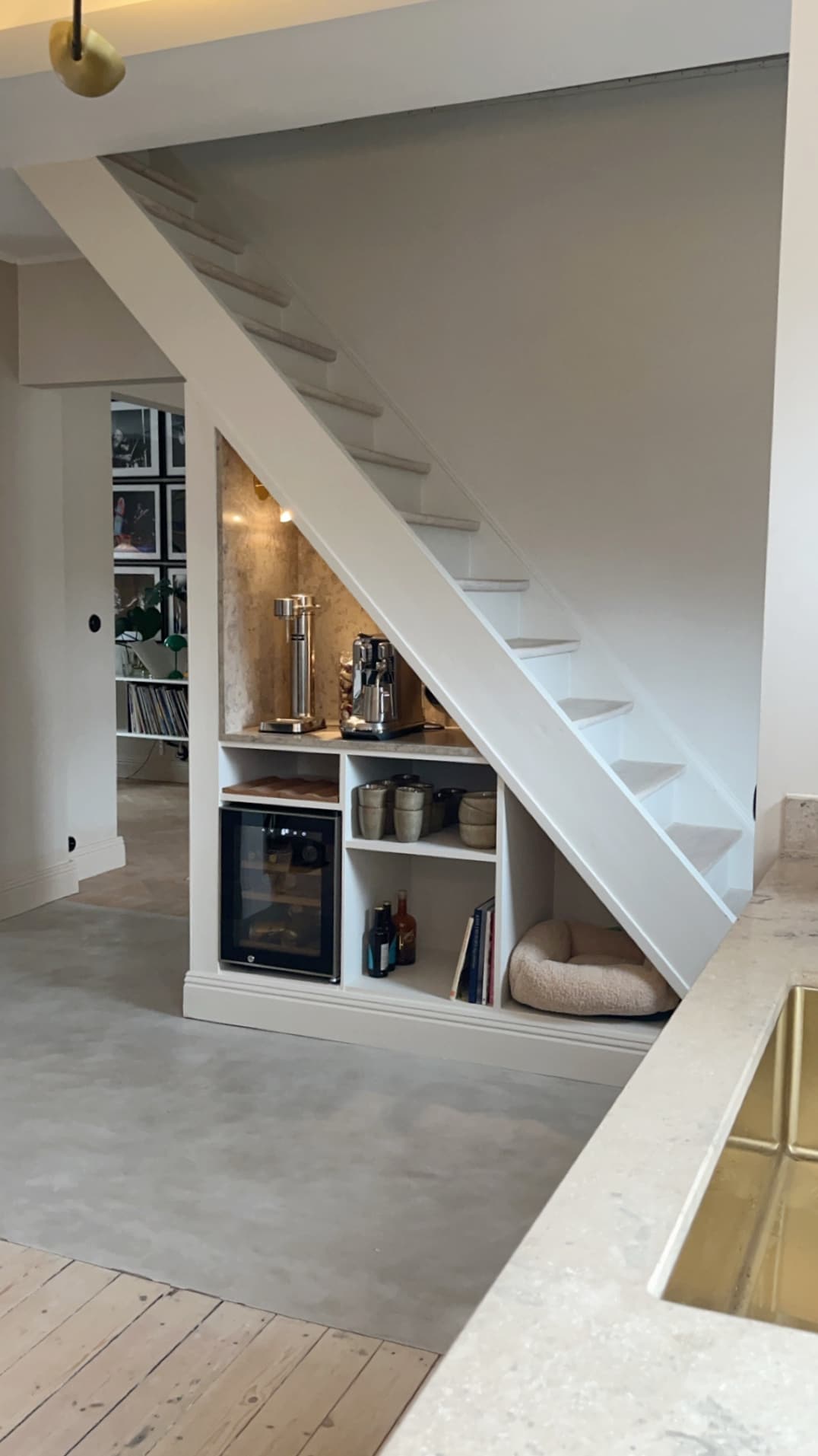 A clean and modern interior features a staircase with white wooden steps. Underneath the stairs, a built-in storage unit displays a wine cooler and decorative items. A cozy pet bed is positioned beside the unit, enhancing the functional space of the home.