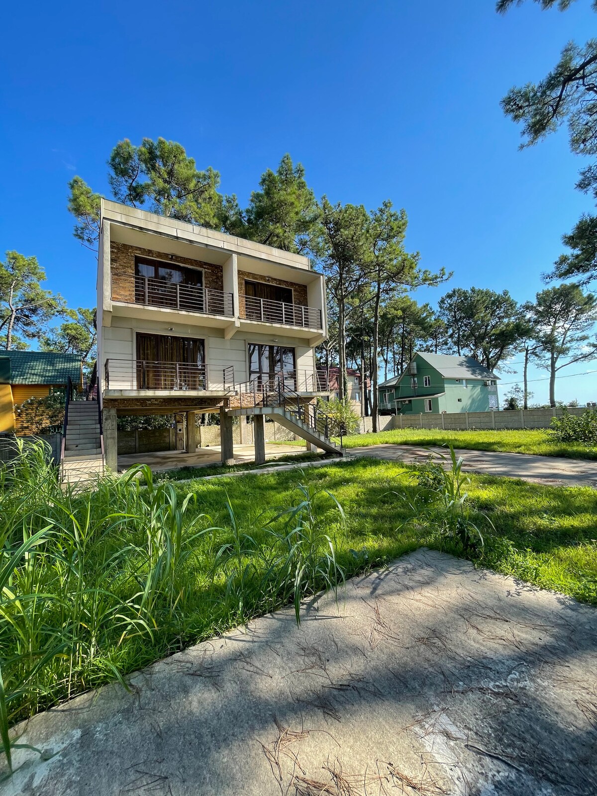 A modern two-story house is framed by tall trees and lush greenery. The exterior features large balconies with wooden slats, providing a connection to nature. A clear blue sky serves as a backdrop, enhancing the tranquil setting. A pathway leads to the entrance, surrounded by grass.