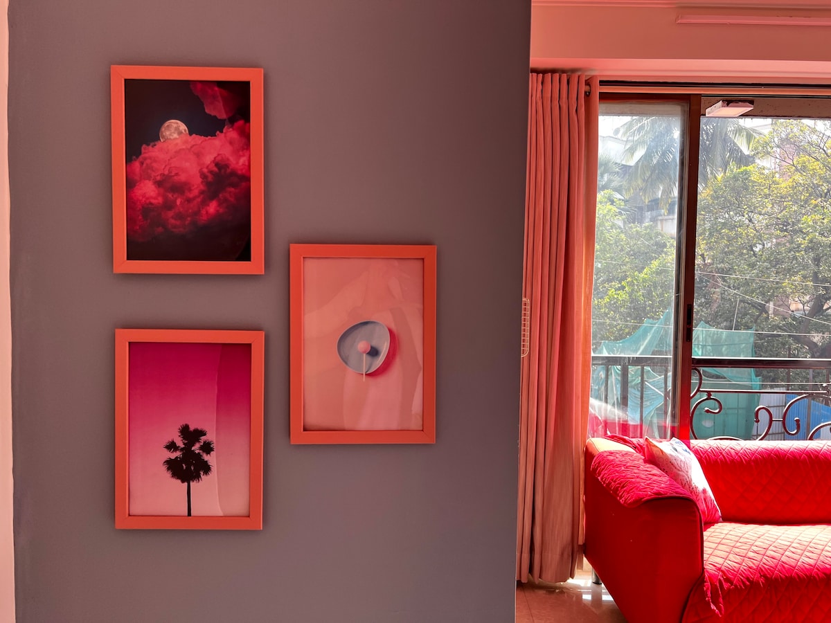 Artfully arranged framed prints adorn the wall, featuring vibrant colors and abstract designs. Natural light streams through the nearby window, highlighting the textures of the fabric sofa and creating a refreshing ambiance in the living space.