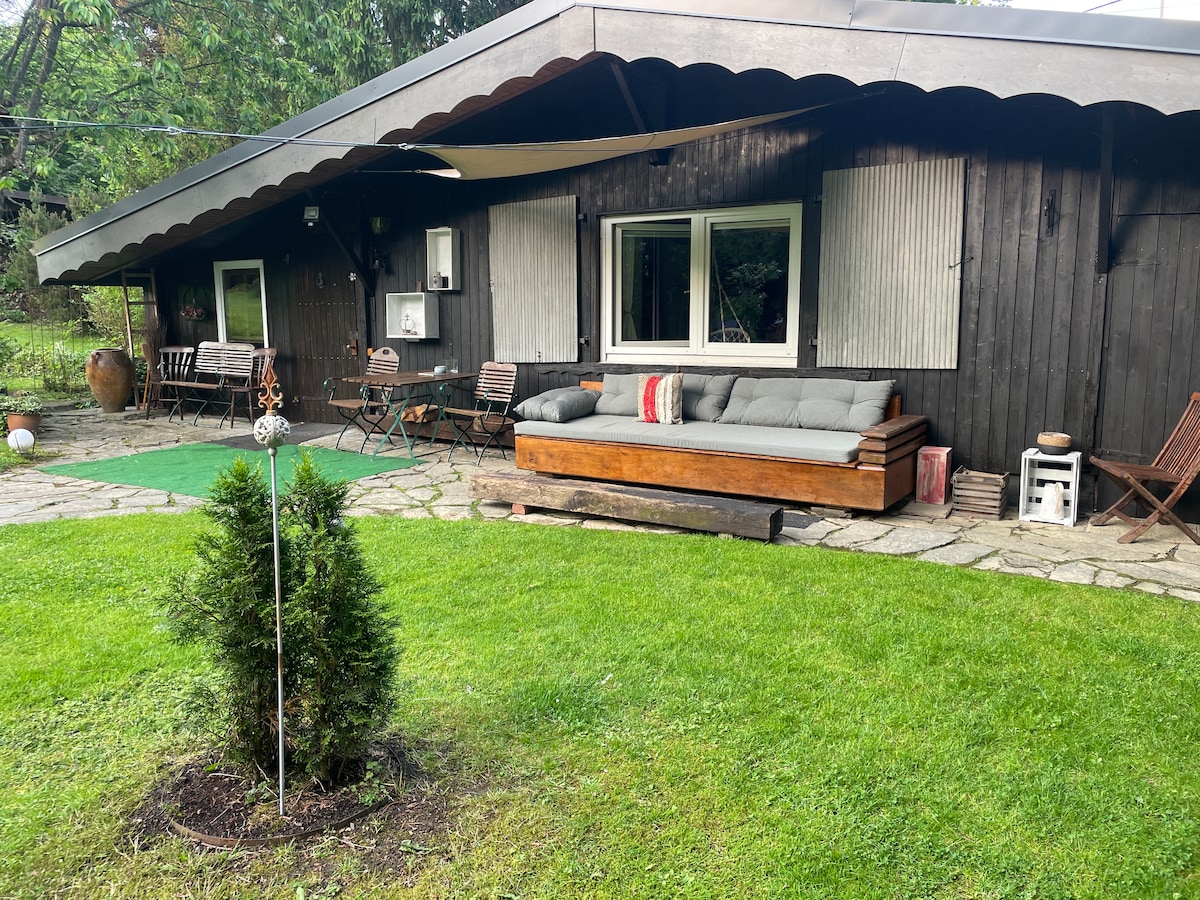 A cozy outdoor seating area is situated in front of a rustic wooden house. Comfortable cushions adorn a long bench, while a small table and chairs are positioned nearby. Lush green grass surrounds the space, complemented by a decorative garden feature.