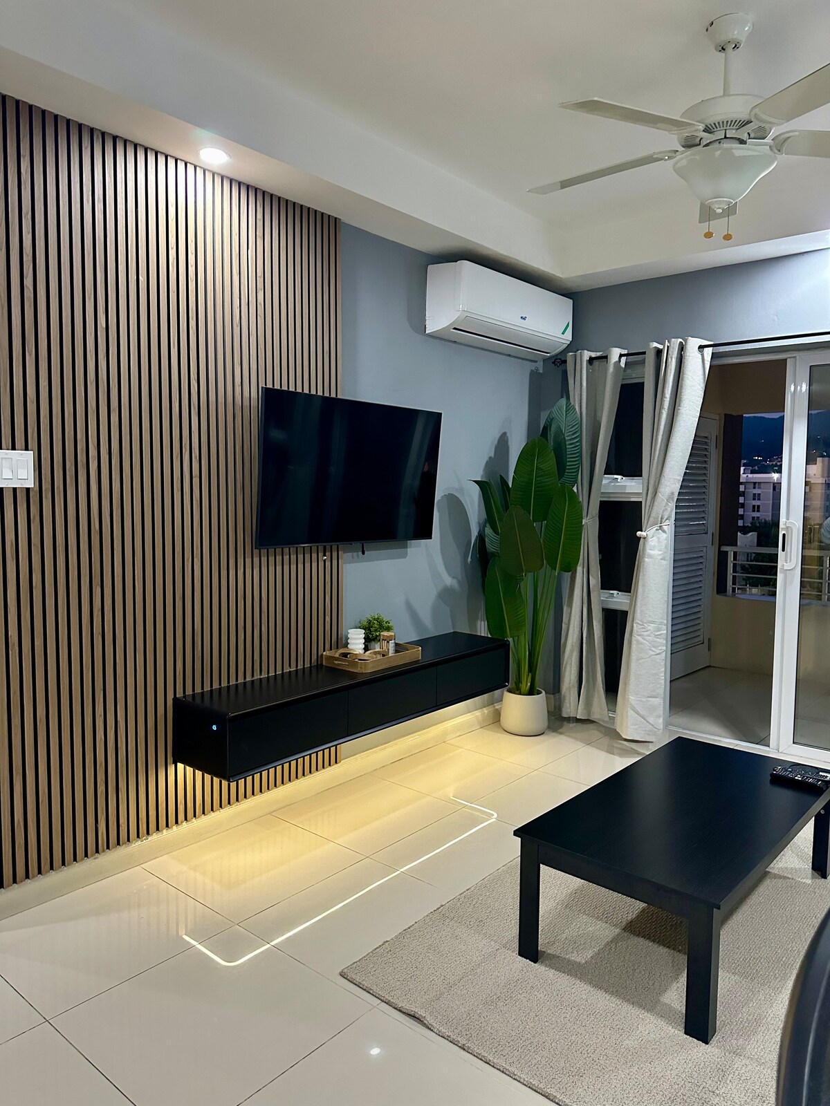 A modern living area features a flat-screen television mounted on a sleek wooden accent wall. A minimalist coffee table sits on a neutral area rug, while a potted plant adds a touch of greenery. Natural light enters through glass doors leading to a balcony.