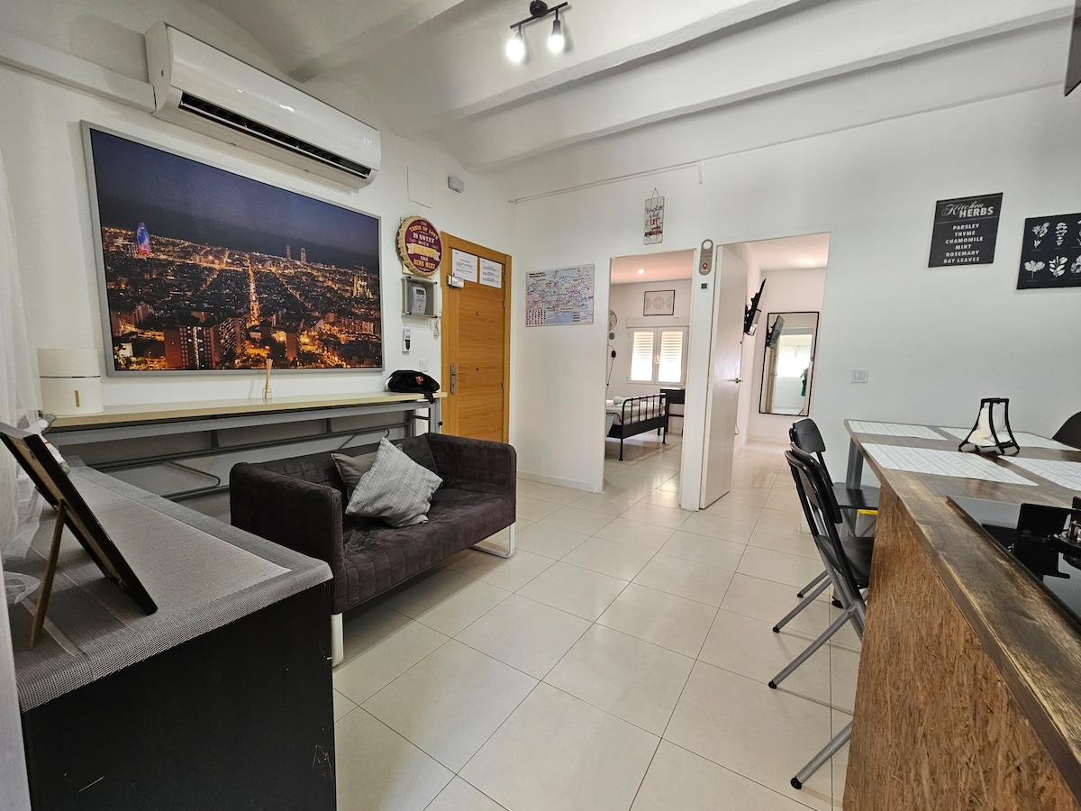 An open living space features a cozy gray sofa accompanied by a decorative pillow. A large wall-mounted air conditioning unit is visible above. A panoramic urban night view adorns the wall, while a wooden counter with seating faces the living area. Natural light illuminates the room.