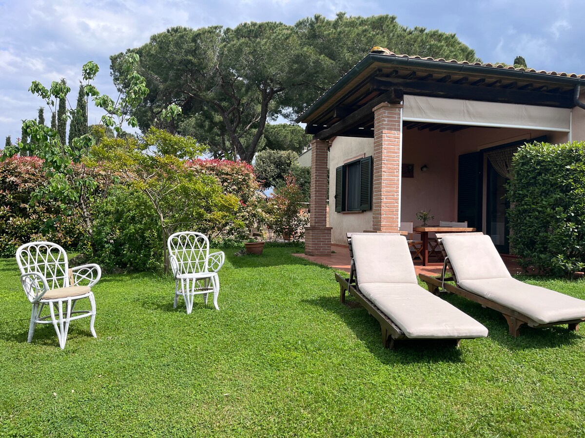 An inviting outdoor space features two sun loungers and two white chairs arranged on well-maintained green grass. In the background, a charming building with a covered porch and dark green shutters complements the surrounding vibrant foliage.