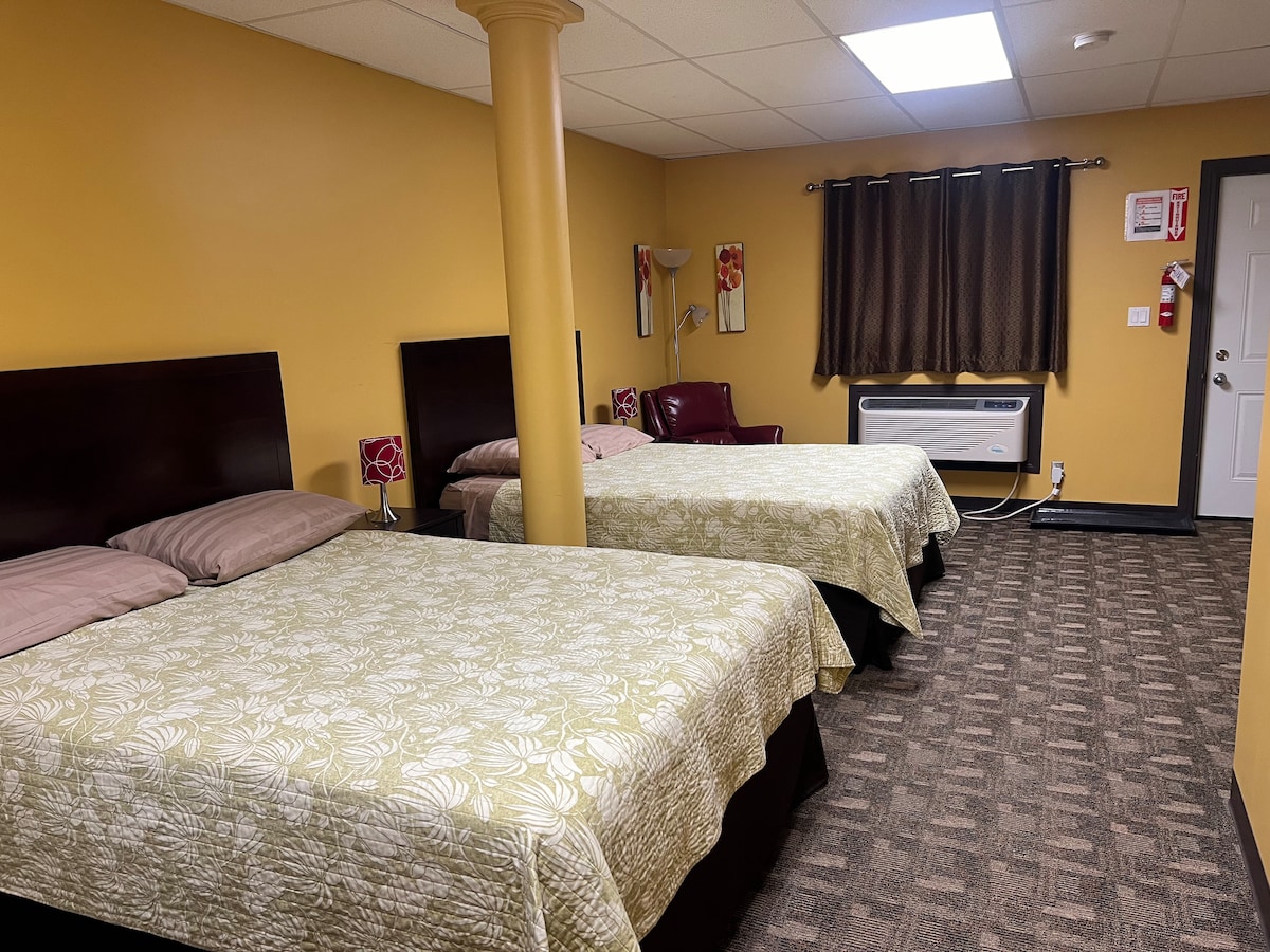 A comfortable sleeping area features two spacious beds adorned with light green floral-patterned bedspreads. A burgundy armchair adds a touch of color, while dark curtains frame the window. A column is visible, along with an air conditioning unit and door leading outside.
