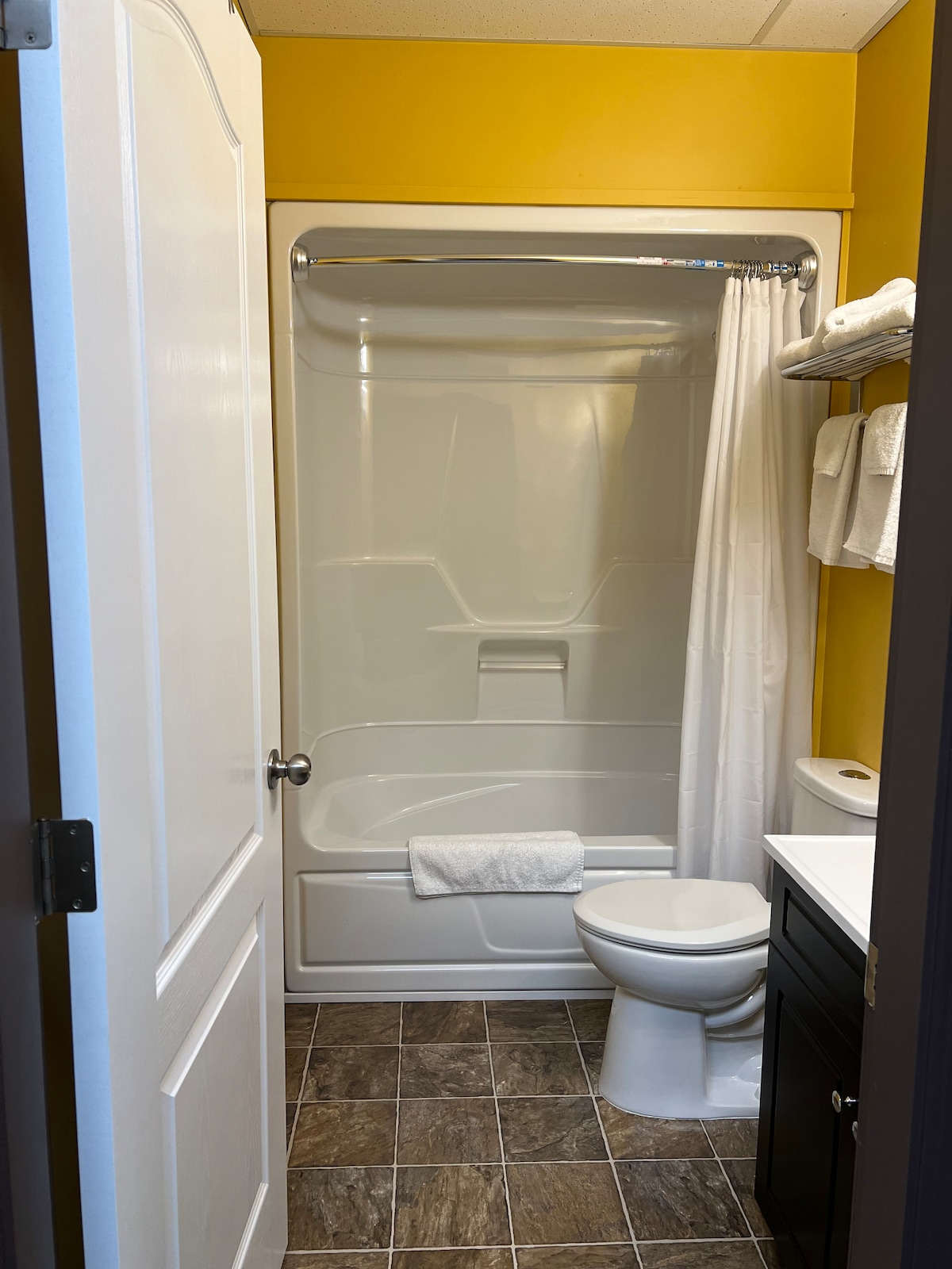 The bathroom features a clean, modern design with a bathtub-shower combination. Soft white towels are neatly arranged on the shelves. Complementary dark cabinetry contrasts with the bright yellow walls, while a light-colored floor adds to the overall brightness.