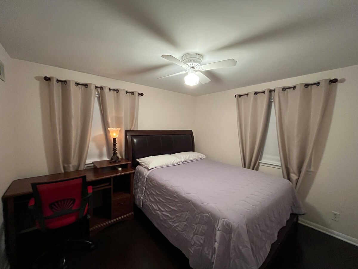 A well-lit bedroom features a spacious bed covered with a light purple quilt. A desk with a red chair is positioned against one wall. Soft curtains frame the windows, and a ceiling fan provides added comfort.