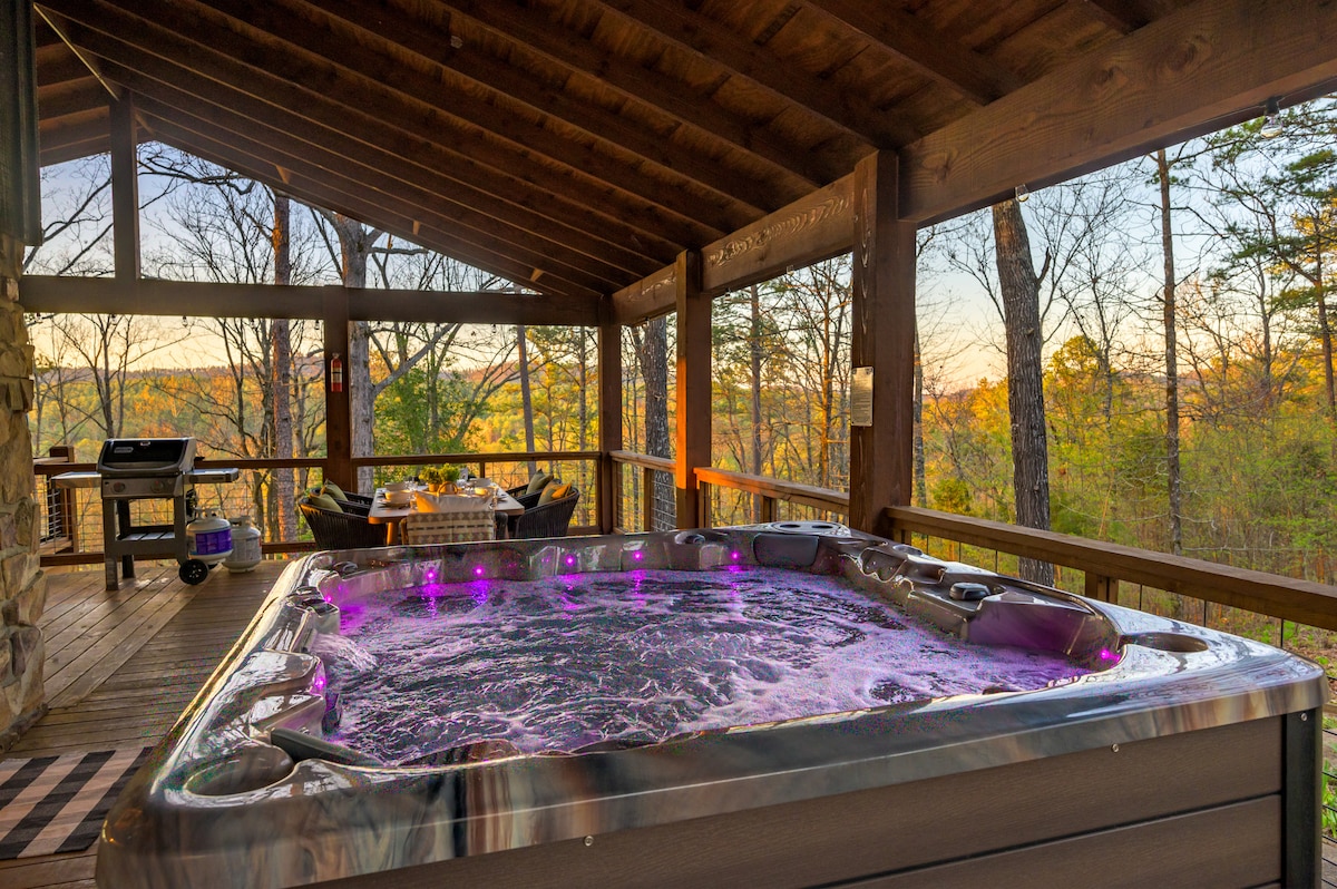 A hot tub with vibrant purple lighting is situated on a covered deck, surrounded by wooden railing that provides an unobstructed view of the forested landscape. A grill and seating area complement the outdoor space, inviting relaxation and enjoyment of the natural surroundings.