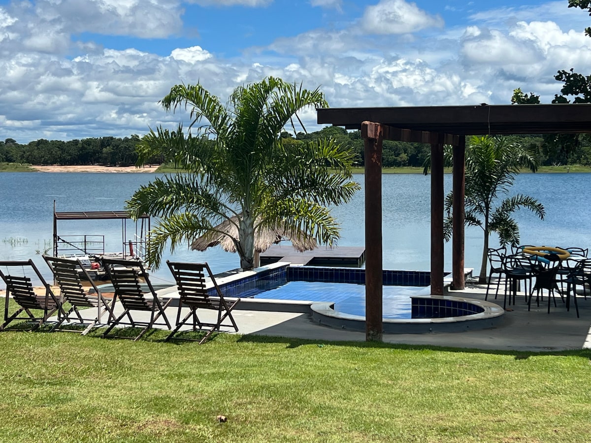 A serene outdoor area features a blue swimming pool accompanied by a separate jacuzzi. Surrounding the pool, wooden lounge chairs are arranged on a green lawn. A shaded patio with dining furniture provides a view of the lake, complemented by lush palm trees.