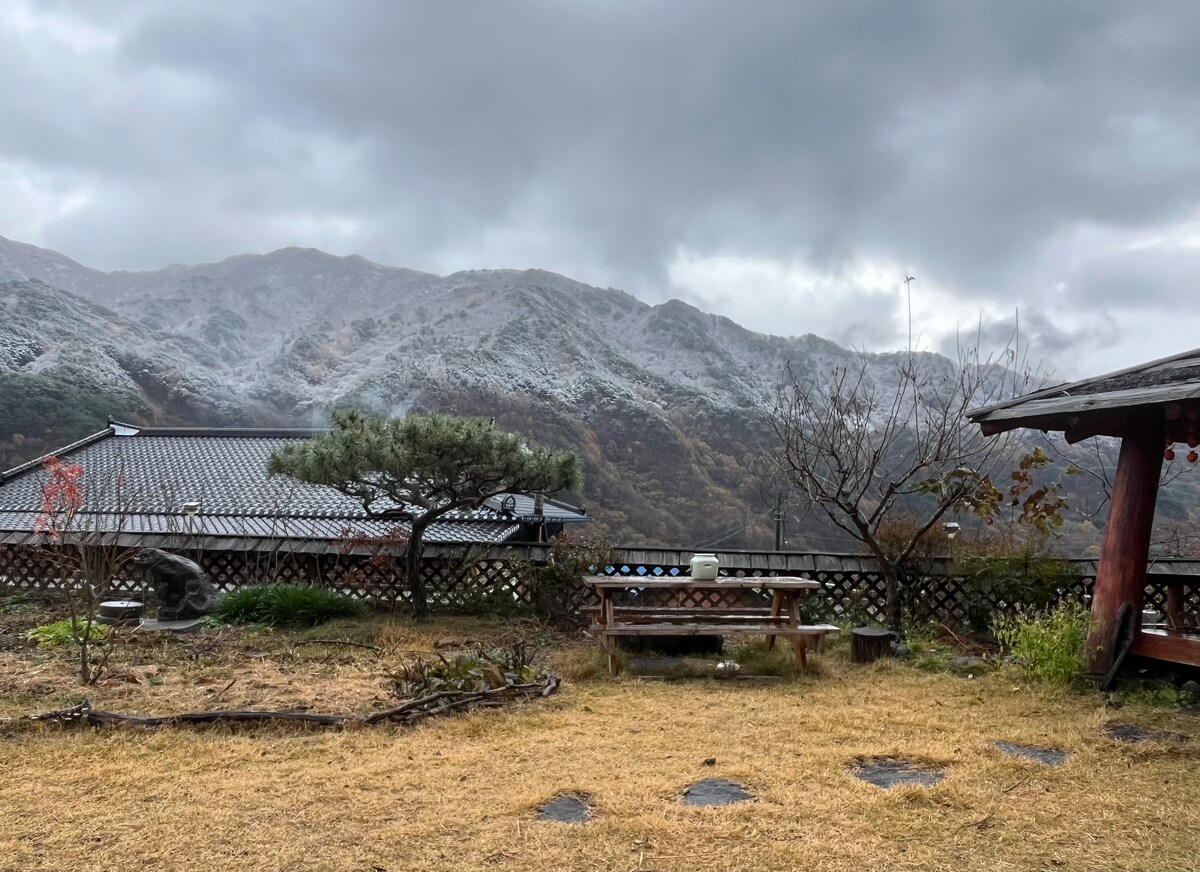 The outdoor area features a grassy space with a wooden picnic table and various plants. In the background, misty mountains capped with snow rise under a cloudy sky, creating a serene and natural landscape. A traditional-style structure is visible on the left.