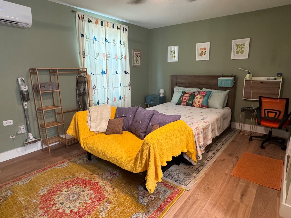 A well-furnished sleeping area features a cozy queen size bed dressed in soft linens with an assortment of colorful pillows. A small desk and chair are located in one corner, while a decorative area rug adds warmth to the wooden floor. Light filters through a curtain adorned with subtle patterns.