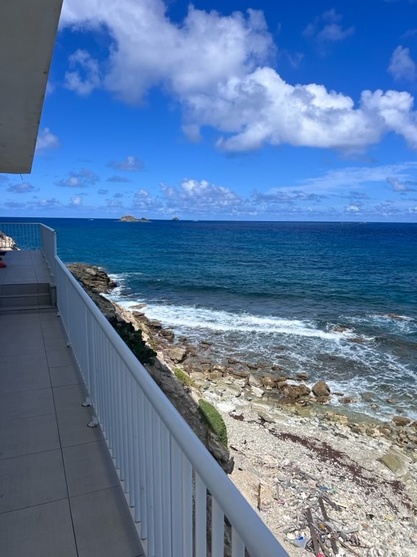 A spacious balcony with a white railing offers unobstructed views of the ocean and rocky shoreline. Gentle waves can be seen lapping against the shore, with a bright blue sky dotted with clouds enhancing the tranquil atmosphere.