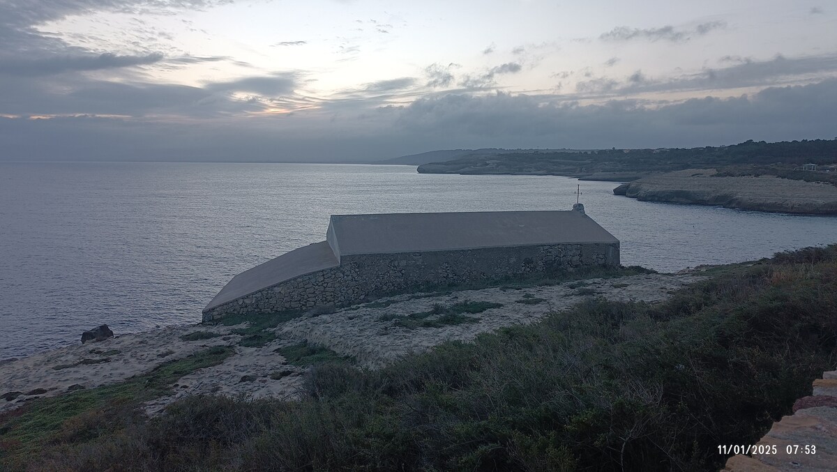 A coastal landscape is depicted, featuring a stone building situated on rocky terrain by the water's edge. The sea extends to the horizon, with gentle waves lapping against the shore. Soft, muted colors characterize the sky, reflecting a tranquil atmosphere during early morning hours.