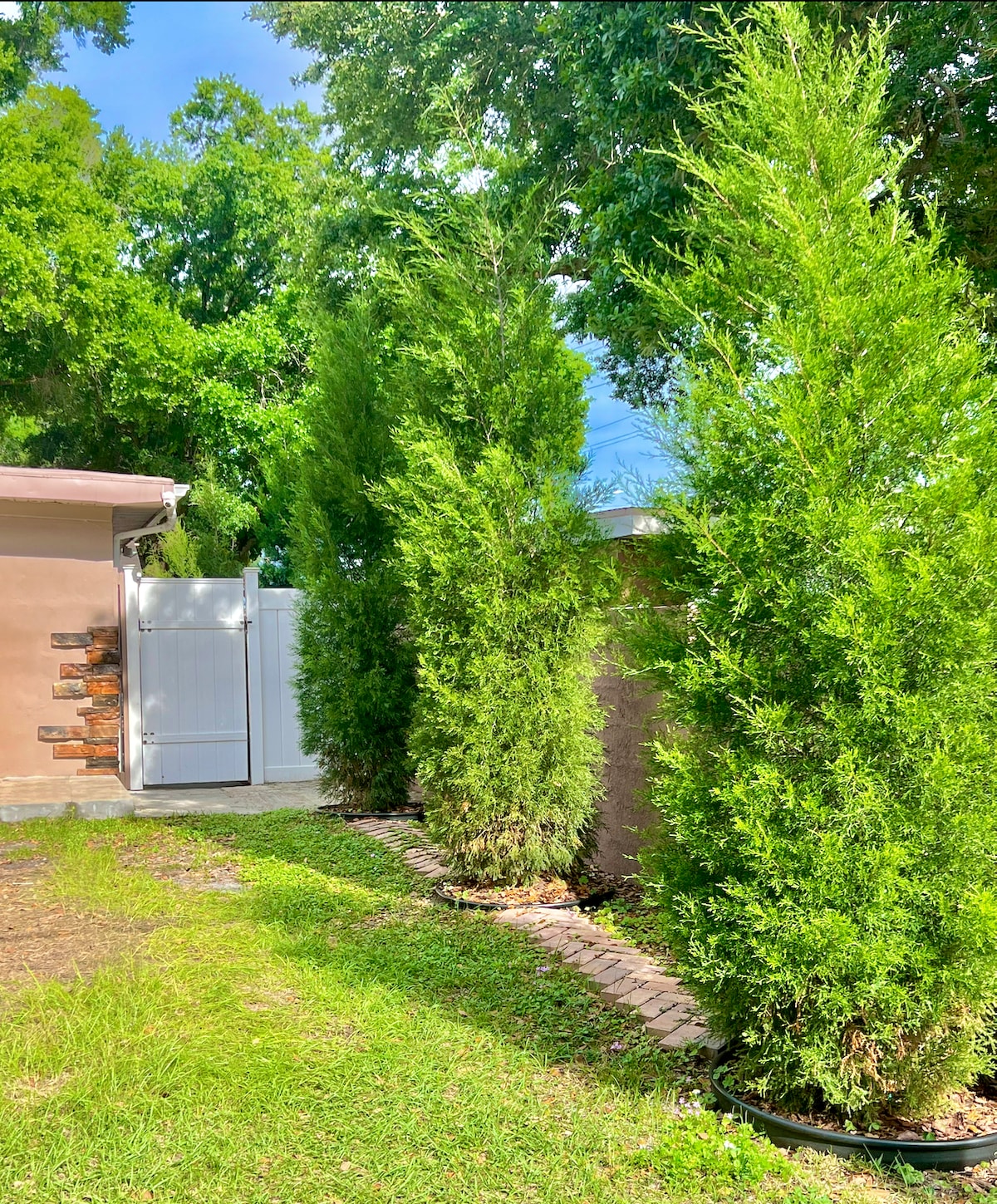 Tall, lush greenery frames the side of the property, with neatly arranged shrubs providing privacy. A white gate is visible at the end of a manicured path, which is bordered by green grass and packed soil.