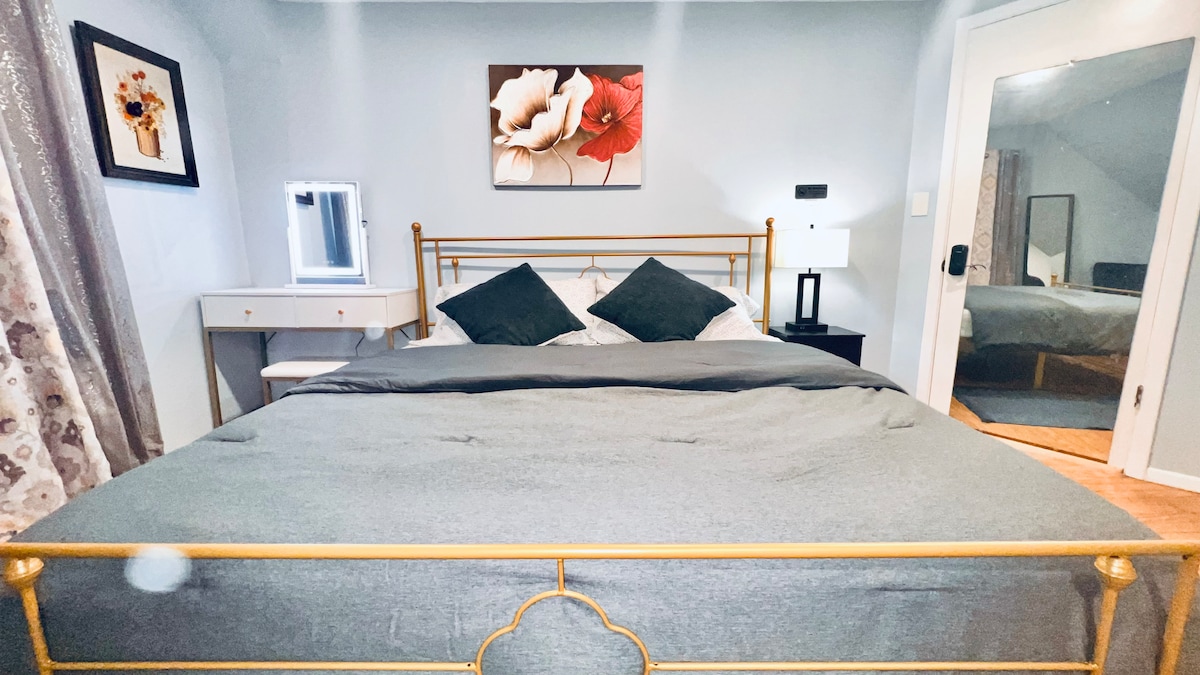 A spacious sleeping area features a gold bed frame with a gray duvet and striped pillows. Soft lighting is provided by a lamp on a nightstand. A decorative mirror and artwork add subtle elegance, while light-colored curtains frame the window, contributing to the overall calming ambiance.