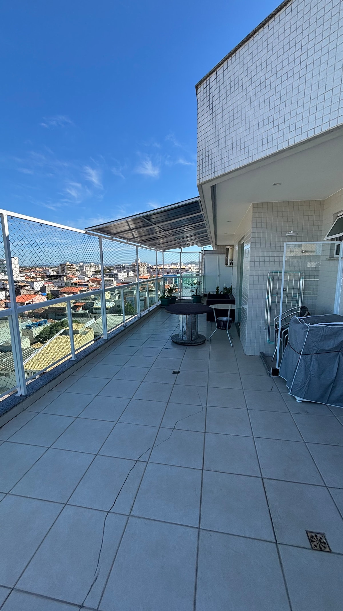 A spacious balcony is visible, featuring tiled flooring and a partial awning overhead. A barbecue grill is positioned at one end, with seating options nearby. The surrounding railing offers views of the cityscape, complemented by potted plants adding greenery to the setting.