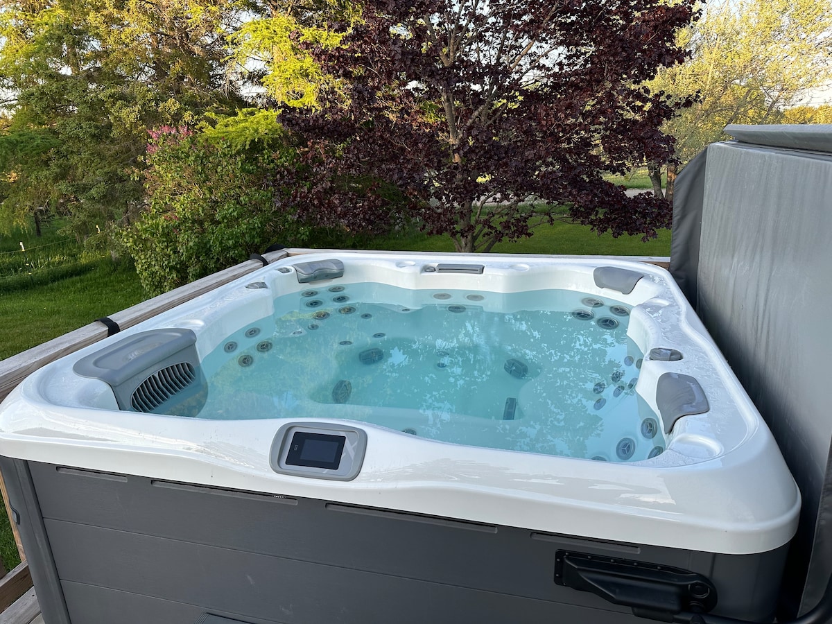 A hot tub sits on a wooden deck, featuring numerous jets and a clear water surface. Surrounding greenery is visible, enhancing the outdoor ambiance. The spa is securely covered on one side, with accessible controls located at the front.