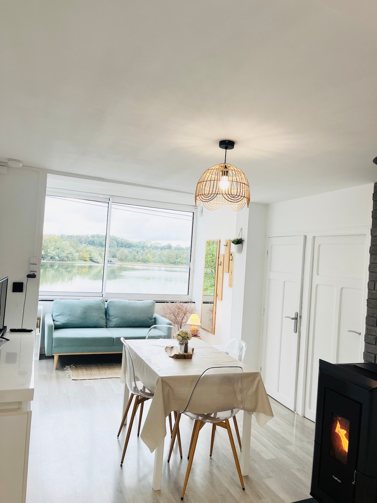 A bright dining area features a light wood table surrounded by four chairs. A soft blue sofa is positioned by large windows offering views of the lake. A cozy fireplace is adjacent, complemented by a woven light fixture above the table.