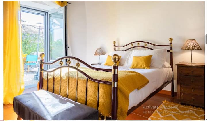 Cama doble luminosa y cómoda con cabecero de felpa, sábanas blancas crujientes y colchas/alfombras acolchadas de color amarillo soleado. Cuenta con dos mesitas de noche con lámparas de ratán y un gran armario de madera para guardar cosas.