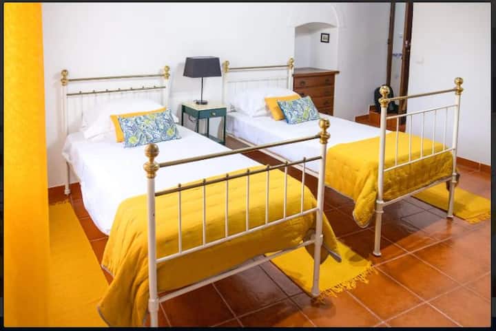 ¡Dos camas individuales te esperan en nuestro dormitorio soleado! Cada una cuenta con sábanas blancas nuevas, una colcha acolchada de color amarillo brillante y almohadas con estampados vibrantes. Una elegante mesita de noche con lámpara y cálidos suelos de baldosas completan este acogedor y confortable espacio.