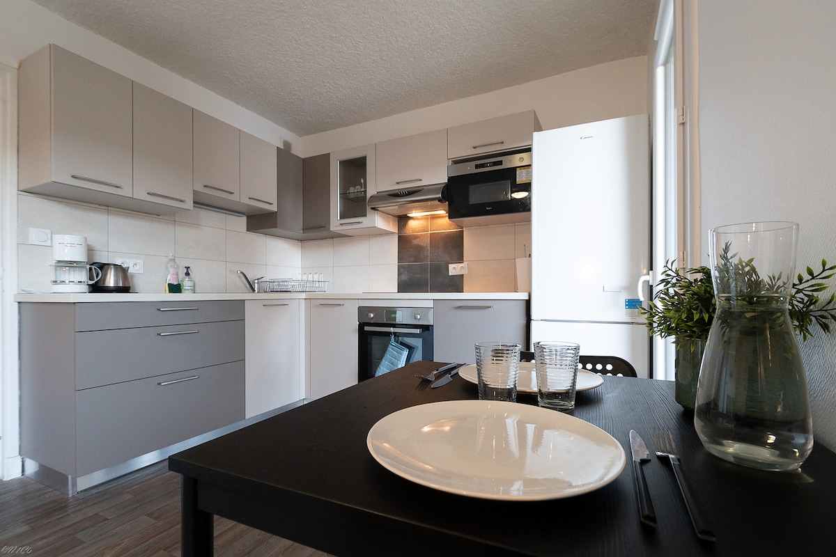 The kitchen showcases modern cabinetry with a minimalist design. It is equipped with essential appliances including an oven, microwave, and refrigerator. A dining table is set for two, featuring plates and glassware, with natural light filtering in from a nearby window.