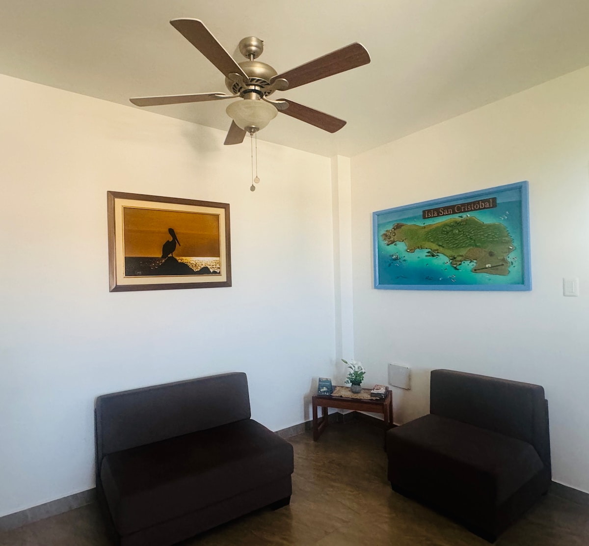 The living area is furnished with two dark sofa beds, one larger than the other. A ceiling fan is positioned above, and artwork depicting a pelican and a map of San Cristóbal adorns the walls. A small table is placed between the sofas.
