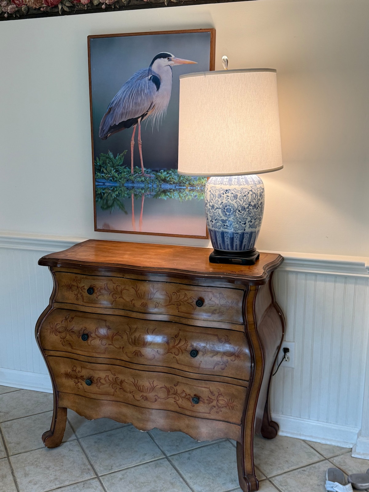 A vintage wooden dresser with decorative carvings stands against a wall, topped by a blue and white lamp. Above the dresser, a framed artwork features a heron amidst greenery, adding a serene touch to the space.